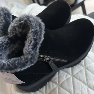 Svarta boots med fuskpäls från Skechers - Svarta boots från Skechers med mjuk grå fuskpäls runt skaftet och dragkedja på sidan. Ovandel i mocka och grov sula för extra grepp. Perfekta för kalla dagar när du vill hålla stilen och värmen.