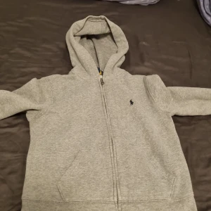 Grå hoodie från Polo Ralph Lauren - Säljer en grå hoodie från Polo Ralph Lauren med dragkedja och klassisk huva. Tröjan har långa ärmar, fickor framtill och den ikoniska Polo-loggan broderad på bröstet. Perfekt för chill dagar och enkel att matcha med jeans eller joggers. Den har två små fläckar vid armen vilket inte syns så jätte mycket. Pris kan diskuteras om du har en minsta fråga bara fråga😊