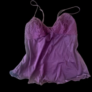 Vinröd linnetopp med tunna axelband - Tröjan är vinröd fast det ser lila ut på bilden! Supersöt lila linnetopp med tunna axelband och plisserad överdel. Toppen har en lätt glansig finish och flowig passform nedtill. Perfekt för dig som vill ha en feminin och trendig look.