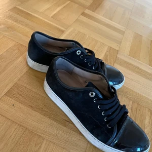 Svarta sneakers från Lanvin med lackad tå - Säljer ett par svarta Lanvin sneakers med snygg lackad tå och mocka på sidorna. Skorna har rund tå, platt vit sula och klassisk snörning. Perfekt för dig som gillar stilrena detaljer och vill ha något som sticker ut lite extra. Bra skick! Storlek 42!
