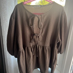 Brun tröja  med snörning från 157 - Säljer en brun tröja  från 157 i storlek S. Klänningen har ett crinkle-texturerat tyg, långa puffärmar och två snörningar framtill vid halsen. Modellen är loose fit med volangdetaljer vid midjan och är perfekt för dig som gillar avslappnad stil.