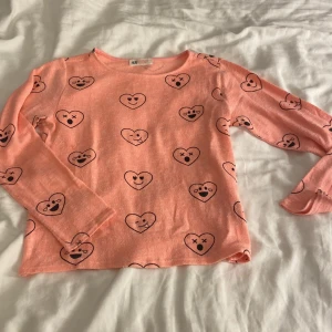 Rosa långärmad tröja med hjärtan H&M - Säljer en söt rosa långärmad tröja från H&M med svarta hjärtan och roliga ansikten som mönster. Tröjan har rund hals och är gjord i mjuk bomull, perfekt för dig som gillar färgglada och lekfulla plagg.