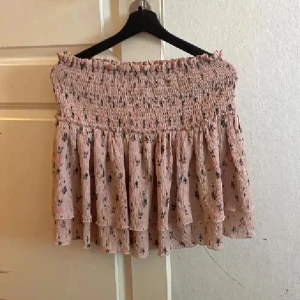 Rosa blommig volangkjol från NA-KD - Supersöt kort kjol från NA-KD i ljusrosa med små blommor. Kjolen har smockad midja och flera lager volanger som ger en flowy och feminin look. Perfekt för dig som gillar romantisk stil och vill ha något luftigt till sommaren.