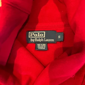 Röd polotröja från Polo Ralph Lauren - Säljer en snygg röd polotröja från Polo Ralph Lauren i storlek S. Tröjan är tillverkad i 100% bomull och har en cool krage med två knappar samt det klassiska blå broderade Polo-logot på bröstet. Perfekt för dig som gillar stilrena och tidlösa plagg.