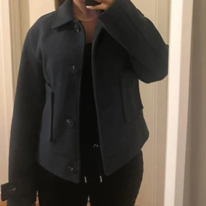 Marinblå ulljacka med knappar - Snygg marinblå jacka i ull med klassisk krage och stora knappar framtill. Jackan har raka, långa ärmar och två fickor framtill. Perfekt för dig som gillar en stilren och enkel look.