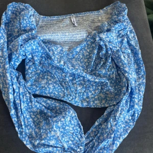 Blommig blå blus med puffärmar - Supersöt blommig blus i blått med vita små blommor. Toppen har v-ringning, smockad rygg och långa puffärmar som ger en romantisk vibe. Perfekt för dig som gillar en feminin och trendig stil.