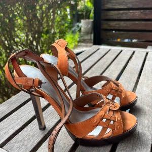 Bruna sandaler med hög klack - Snygga bruna sandaler från French Connection i mocka med flätade remmar och nitar. Skorna har öppen tå, hög klack och justerbar ankelrem. Perfekta för dig som vill sticka ut med en chic och trendig look.