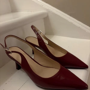 Vinröda pumps med spetsig tå - Snygga vinröda pumps med lackad finish och spetsig tå. Skorna har en låg klack och öppen häl med justerbart spänne. Perfekta för dig som vill sticka ut med en elegant och trendig look.