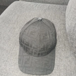 Svart Fendi keps med logomönster - Snygg svart keps från Fendi med klassiskt FF-logomönster över hela kepsen. Justerbar rem baktill med Fendi-tryck och böjd skärm. Tillverkad i ett tygmaterial som känns lyxigt och stilrent, perfekt för dig som vill sticka ut med designeraccessoar.