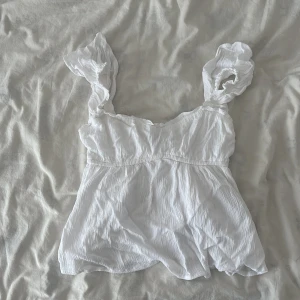 Vit volangblus från Hollister - Supersöt vit blus från Hollister med volangiga axelband och lätt rynkad struktur. Toppen har en feminin v-ringning och smockad midja som ger en snygg passform. Perfekt till jeans eller kjol för en fräsch look. Någon storlek för liten på mig(168 cm)