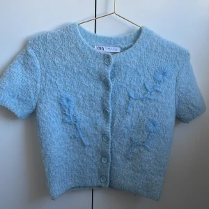 Ljusblå stickad kofta från Zara - Superfin ljusblå stickad kofta från Zara med korta ärmar och rund hals. Koftan har knappar framtill och broderade blommor i samma färg som ger en söt vibe. Perfekt att styla med jeans eller kjol för en chill look.