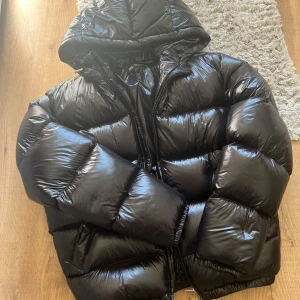 Svart pufferjacka från ZARA med huva - Säljer en svart, glansig pufferjacka från ZARA med huva och dragkedja. Jackan är vadderad och har en oversized look, perfekt för kalla dagar. Materialet är syntet och jackan har långa ärmar samt praktiska fickor på sidorna. Använd endast någon gång då det visade sig inte vara min smak. 