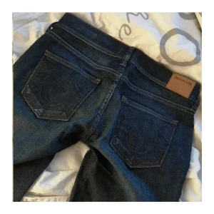 True Religion Cora Straight jeans blå - Snygga mörkblå jeans från True Religion, modell Cora Straight. Klassisk femficksdesign med raka ben och ikoniska bakfickor. Jeansen har en stilren look och är tillverkade i slitstarkt denim med snygga detaljer och logopatch bak.