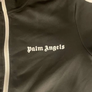 Svart Palm Angels zip hoodie - Svart zip hoodie från Palm Angels med vita ränder längs ärmarna och logga tryckt på bröstet. Tröjan har lång ärm, ståkrage och dragkedja framtill. Materialet är mjuk polyester som ger en sportig vibe. Perfekt för dig som gillar streetwear och snygga detaljer.