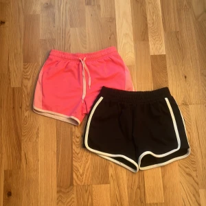 Två sportiga shorts med kontrastkant - Säljer två par sportiga shorts, ett par i stark rosa med ljusrosa kant och snörning i midjan, samt ett par svarta med vit kant. Båda har elastisk midja och är tillverkade i ett lätt, ventilerande syntetmaterial. Perfekta för träning eller chill på sommaren.svarta:20kr storlek 146,rosa:15kr eftersom det är lite fläckar där bak storlek 122/128,båda:30kr