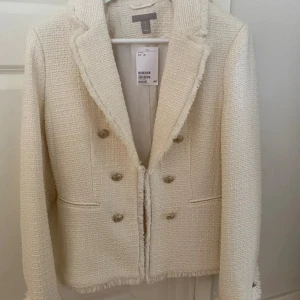 Beige kavaj från H&M med fransdetaljer - Snygg beige kavaj från H&M med dubbelknäppning och dekorativa guldknappar. Jackan har fransade kanter och är tillverkad i ett strukturerat tyg som ger en exklusiv känsla. Perfekt för dig som vill ha en stilren och trendig look.