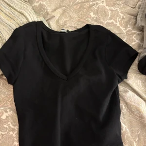 Svart v-ringad topp med kort ärm - En svart, enkel topp med v-ringning och korta ärmar. Perfekt att matcha med jeans eller kjol för en clean look. Gjord i mjukt bomullsmaterial som känns skönt mot huden.