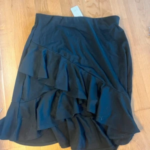 Svart volangkjol med asymmetrisk kant - Supersnygg svart kortkjol från NA-KD med volanger och asymmetrisk nederkant. Kjolen har en clean look och är gjord i mjukt bomullstyg som sitter bekvämt. Perfekt för dig som gillar att sticka ut med coola detaljer och volanger. Nypris  449 och aldrig använd 