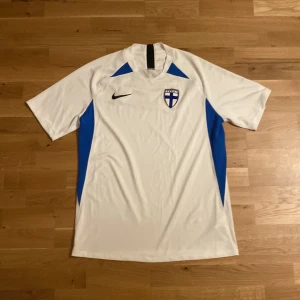 Vit-blå Finland Nike fotbollströja L - Säljer en vit och blå Finland fotbollströja från Nike i storlek L. Tröjan har korta ärmar, Dri-Fit material som andas och är lätt. Finska landslagets emblem på bröstet och svart Nike-logga. Snygga blå paneler längs sidorna för extra stil. Tröjan har en jätte liten fläck som inte ens syns när man har på sig den. Använd max 2 gånger av mig. Kommer från ett mystery football shirt paket från pardon my kicks i höstas/vintras ungefär 