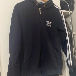 Svart Adidas hoodiejacka med dragkedja - Svart hoodiejacka från Adidas med vit logga på bröstet och klassiska vita ränder längs ärmarna. Jackan har huva med snörning, hel dragkedja framtill och fickor med dragkedja. Perfekt för en sportig och avslappnad stil.