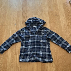 Blårutig skjorta med huva från Zara - Snygg blårutig skjorta från Zara med huva och knappar framtill. Skjortan har lång ärm och ett klassiskt rutmönster i blått, vitt och grått. Perfekt för dig som gillar avslappnad stil och vill ha något som sticker ut lite extra.