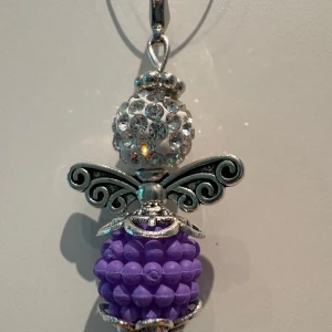 Lila och silver ängelhalsband - Unikt halsband med en ängelberlock i silverfärgad metall. Kroppen är en lila pärla med bubblig struktur och huvudet är en glittrig pärla med strass. Vingarna har snygga snirkliga detaljer. Perfekt för dig som gillar personliga smycken med lite extra bling.
