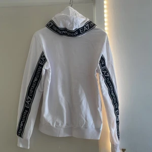 Vit EA7 hoodie med logoband (oanvänd) - Säljer en vit ea7/ Armani hoodie som jag fick på julen men har tyvärr inte kommit till andväning, hoddien är helt oanvänd samt inga fläckar eller skador. Ord priset på hoodien är 1722kr. Hoodien har text ränder på armarna och huvan och är en stretchig hoodie. Passar bra nu när de börjar bli lite kallare ute.