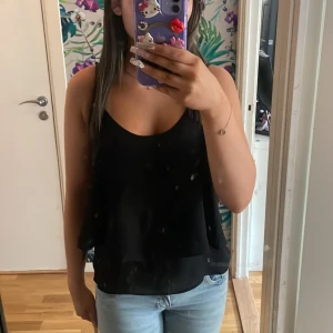 Svart flowy linnetopp med smala band - Säljer en svart, luftig linnetopp med smala axelband och snyggt korsad detalj i ryggen. Toppen har ett flowy lager och är tillverkad i ett lätt, transparent material som ger en chill vibe. Perfekt för varma dagar och enkel att styla med jeans eller shorts.