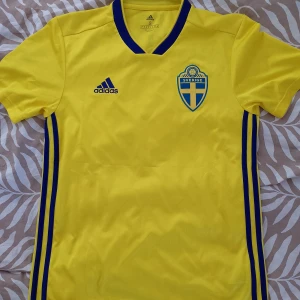 Svensk landslagströja Adidas S - Gul fotbollströja från Adidas med blå detaljer, officiell Sverige-emblem på bröstet och tryck WALKER 9 på ryggen. V-ringad krage med blå kant, korta ärmar och Climalite-material som andas. Perfekt för dig som vill representera Sverige på match eller träning. Se bilder för storlek och skick. 