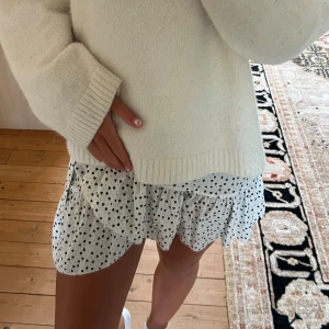 Vit kortkjol med svarta hjärtan - Supersöt vit kortkjol med svarta hjärtan som mönster. Kjolen har volangkant och en luftig, flowy passform som ger en skön och lekfull vibe.💛