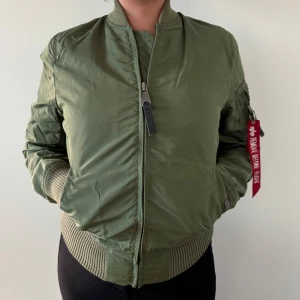 Grön bomberjacka från Alpha Industries - Säljer en klassisk grön bomberjacka från Alpha Industries med orange foder och ikonisk 'Remove Before Flight'-tag på ärmen. Jackan har dragkedja, ribbade muddar och flera fickor. Perfekt för dig som gillar streetwear och tidlös stil.