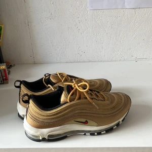 Nike Air Max 97 Gold sneakers - Nike Air Max 97 sneakers i metallic guld med svarta och vita detaljer, röd swoosh på sidan och synlig Air-sula. Ovandelen har vågiga linjer och snörning i matchande guld. Skorna har en platt sula och rund tå, samt dragflik med Air Max-tryck bak. Perfekt för dig som gillar att sticka ut. Sparsamt använda 4-5 gånger. Säljes utan box. Köpta från sneakersnstuff. 