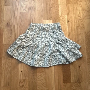 Grå blommig volangkjol från 157 New - Supersöt kort kjol med volanger från 157 New. Kjolen är grå med ett blommigt mönster i vitt och svarta detaljer. perfekt för dig som gillar en chill och lekfull stil.