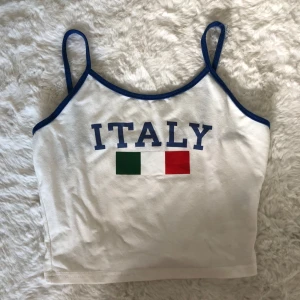Vit croppad linne-topp ITALY -  en vit croppad linne med blå axelband. Framtill står det ITALY i blått med italienska flaggan under. På ena axelbandet så har sömmen gått sönder lite (vilket man kan fixa enkelt).