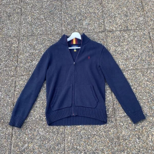 Mörkblå stickad kofta från Polo Ralph Lauren - Snygg mörkblå stickad kofta från Polo Ralph Lauren i storlek XL. Tröjan har dragkedja framtill, ribbade muddar och fickor. Kragen har coola ränder i vinrött och gult. Perfekt för dig som gillar klassisk stil med en twist. Det är xl i barnstorlek och är då i storlek 176 cm också!