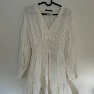 Vit klänning med v-ringning från Zara - Supersöt vit klänning från Zara med v-ringad hals och långärmade ärmar. Smockad midja och ärmslut ger en snygg siluett och volang nedtill gör den extra charmig. Lite tjockare material i linne. Användbar både under sommar och vinter.