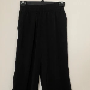 Svarta wide byxor från H&M Divided - Svarta satin liknande byxor från H&M Divided med breda ben och resår i midjan. Tillverkade i 100% polyester för en lätt och luftig känsla. Perfekta för dig som gillar en avslappnad och trendig stil.