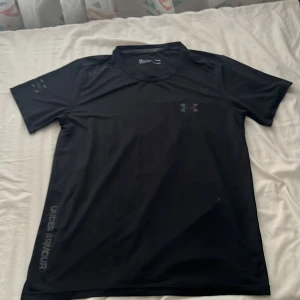 Svart Under Armour träningst-shirt - Svart Under Armour t-shirt i funktionsmaterial med korta ärmar och reflekterande logga på bröstet. Snygga detaljer med UA-SPEC-tryck på axeln och Under Armour-tryck längs sidan. Perfekt för dig som gillar sportig och clean stil.
