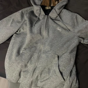 Burberry hoodie grå - Grå hoodiejacka från Burberry med dragkedja, snörning i huvan och klassiskt rutigt foder på insidan. Jackan har en broderad logga på bröstet och stora fickor framtill. Perfekt för dig som vill ha en stilren men ändå lyxig look.