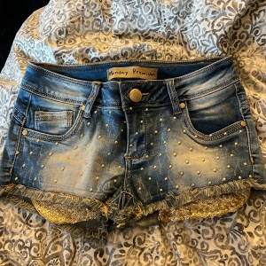 Blå jeansshorts med pärlor från Monday  Premium - Snygga blå jeansshorts från Monday Premium i storlek S. Shortsen har guldiga pärlor på fram- och bakfickor, fransiga kanter och glittriga paljettdetaljer vid bensluten. Klassisk femficksmodell med knapp och dragkedja framtill. Perfekt för dig som gillar att sticka ut.