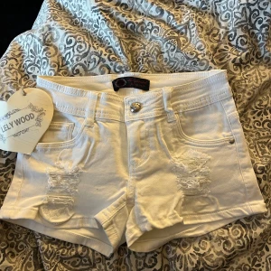 Vita jeansshorts från Lelywood - Snygga vita jeansshorts från Lelywood med hög midja och slitna detaljer framtill. Klassisk femficksmodell med knapp och dragkedja. Perfekta för sommarens alla äventyr, med en fräsch och clean look som passar till allt.