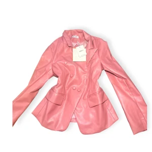 Rosa skinnkavaj med knappar på engelska - Snygg rosa kavaj i skinnimitation med dubbelknäppning och klassisk krage. Jackan har långa ärmar, två fickor framtill och en figurnära passform. Perfekt för dig som vill sticka ut med en färgstark look.