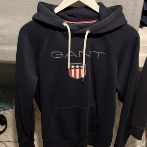 Marinblå hoodie från GANT med logga - Säljer en marinblå hoodie från GANT i storlek M. Tröjan har broderad GANT-logga och sköld på bröstet, vit snörning i huvan och en stor magficka. Materialet är mjuk bomull och polyester, perfekt för chill dagar. Snygga detaljer med metalländar på snöret.
