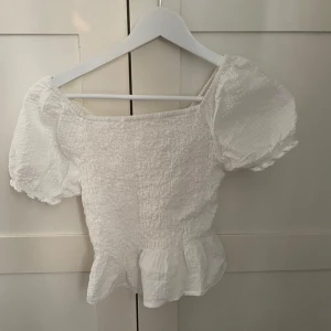 Vit smockad blus med puffärm - Supersöt vit blus med smockad kropp, puffiga korta ärmar och volang nertill. Offshoulder-modell med elastisk halsringning och lätt struktur i tyget. Perfekt för en fräsch och trendig look.