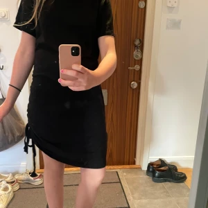 Svart kortklänning med snörning - Säljer en svart kortklänning med snörning i sidan. Klänningen har korta ärmar och en enkel, rak passform som sitter snyggt på kroppen. Perfekt för dig som gillar en stilren look med en liten twist. Materialet känns mjukt och bekvämt.
