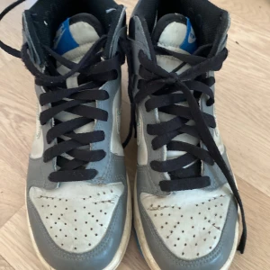 Nike Dunk High grå/vit med blå sula - Nike Dunk High sneakers i grått och vitt med svart snörning och blå yttersula. Klassisk perforerad tå och högt skaft. Ovandel i skinn och syntet, ikonisk Nike-logga på sidan. Perfekta för dig som gillar retrostil och streetwear.