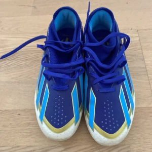 Adidas Messi blå fotbollsskor använt 1 gång och mycket bra skick  - Snygga Adidas Messi fotbollsskor i blått med guld- och turkosa detaljer. Skorna har snörning, platt sula med dobbar och är tillverkade i syntetmaterial. Perfekta för dig som vill sticka ut på planen med stilrena och sportiga skor inspirerade av Messi.