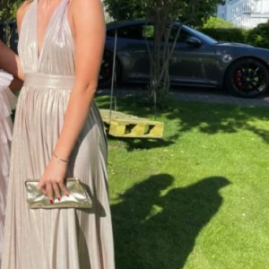 Champagnefärgad långklänning med v-ringning - Säljer en elegant långklänning i champagnefärg med djup v-ringning och bred midja. Klänningen har plisserat tyg som ger ett lyxigt fall och är ärmlös med smala axelband. Perfekt för bal eller festliga tillfällen.
