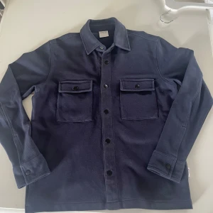 Mörkblå overshirt från Selected Homme - Snygg mörkblå overshirt från Selected Homme med klassisk krage och två stora bröstfickor med knappar. Jackan har raka linjer, knäppning framtill och är tillverkad i ett kraftigare bomullstyg. Riktigt snygg med en vit tisha under! Overshirten kan också användas som ”jacka” under sommarn då den har perfekt tjocklek för det. Tröjan lutar långt mot Stockholmsstil/stekar hållet. 
