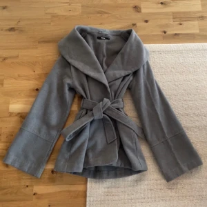 Grå omlottkappa från Bikbok - Snygg grå kappa från Bikbok i omlottmodell med bred krage och bälte i midjan. Jackan har långa ärmar och är tillverkad i ett mjukt ull-liknande material. Perfekt för dig som vill ha en stilren och trendig look💕 Passar till våren, hösten eller kallare sommardagar och kvällar